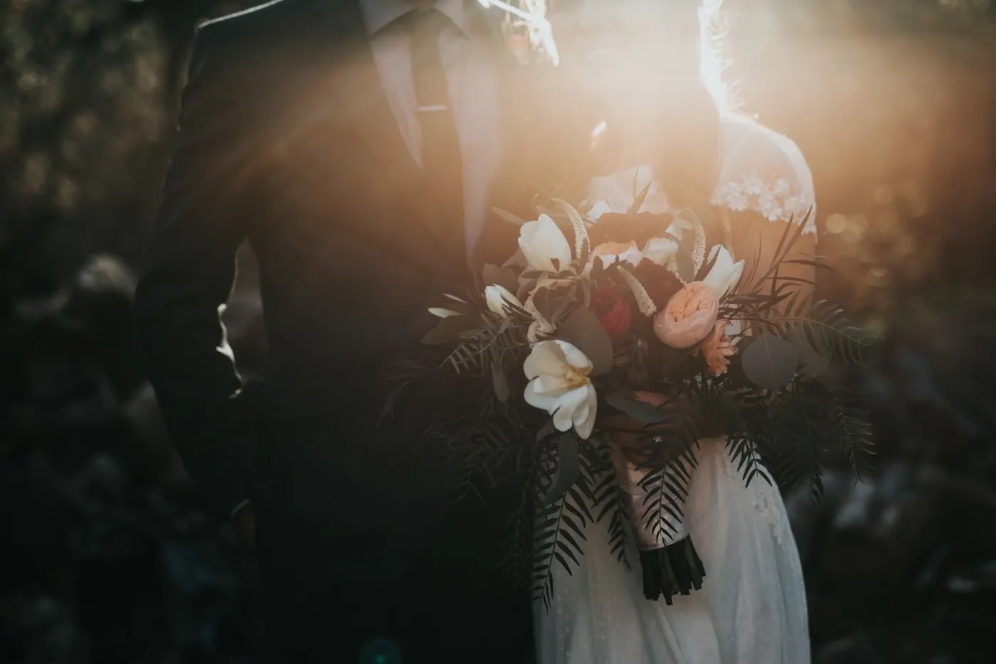 Elena and Marcus close together outdoors, golden backlight through the trees, Elena holding her bouquet
